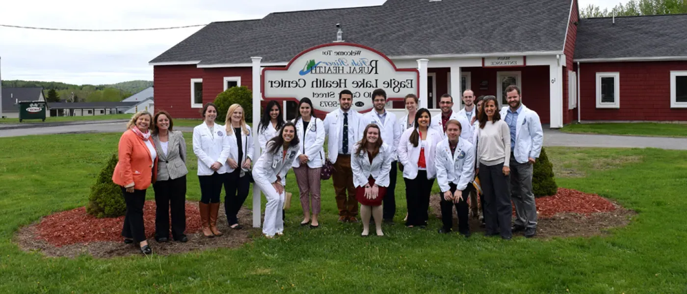 A group of U N E Health professions students pose in white coats in front of the sign for Fish River Rural Health Center